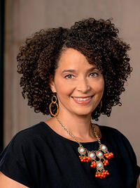 Portrait of Erica Edwards, a light-skinned Black woman with curly hair, wearing a black top adorned with a bright orange and turquoise necklace, in front of a stone wall.